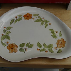 May include: A white ceramic serving platter with a scalloped edge and a brown rim. The platter is decorated with a floral design of orange flowers and green leaves.