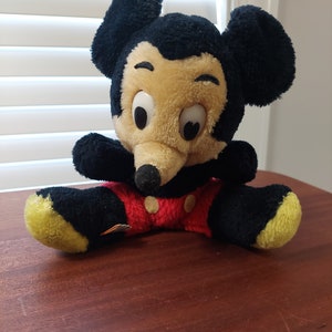 May include: A plush black and yellow Mickey Mouse toy wearing red shorts with white buttons. The toy is sitting on a wooden surface.