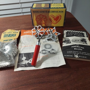 May include: A Nordic Ware double rosette and timbale iron set with a red handle, a cardboard box labeled Double Rosette and Timbale Iron, a package of tart shell molds, a Scandinavian recipe sheet, and an Unusual Old World and American Recipes booklet.