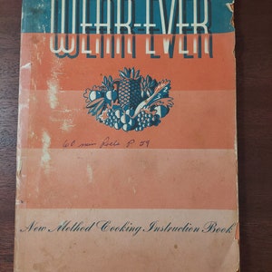 May include: A vintage cookbook with a blue and orange cover featuring the text "WEAR-EVER ALUMINUM" and a colorful illustration of fruit. The text "New Method Cooking Instruction Book" is printed at the bottom of the cover.