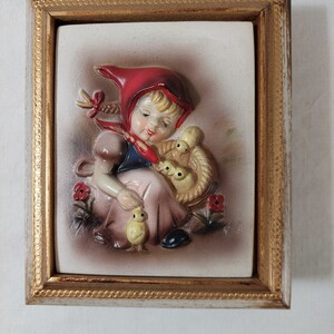 May include: A framed ceramic wall plaque featuring a girl in a red hooded dress holding a basket of chicks. The plaque has a 3D effect and is painted in a vintage style.