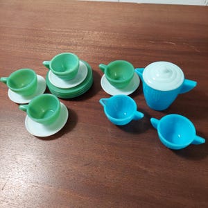 May include: A vintage children's tea set with green and blue cups, saucers, a teapot, and a creamer. The set is made of glass and has a white rim.