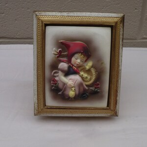 May include: A framed ceramic wall plaque featuring a girl in a red hooded dress holding two yellow chicks. The plaque has a white background and is surrounded by a gold frame.