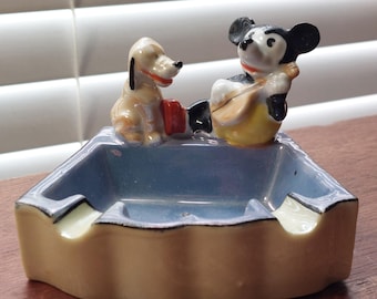 May include: Vintage ceramic ashtray featuring Mickey Mouse playing a lute and Pluto. The ashtray has a light blue interior and a beige exterior with black trim. A collectible Disney item.