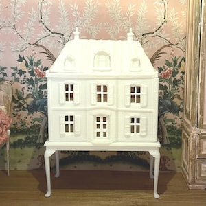 May include: A white dollhouse with six windows and a pitched roof, set on a matching white stand with curved legs. The dollhouse has a classic design and is placed in front of a pink floral wallpaper.