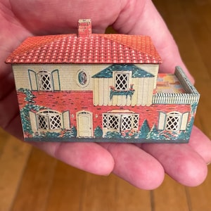 May include: A miniature toy house with a red tiled roof and a chimney. The exterior features a brick-patterned lower level, cream-coloured upper walls, lattice-style windows with shutters, a central door, and printed greenery along the base.