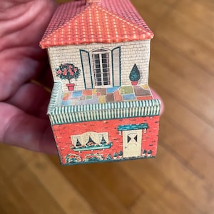 May include: A miniature paper house model featuring a red tiled roof, a stone-textured upper level with a central door, and a lower level with red brick walls. The design includes a multi-coloured patio, potted plants, and a window with flower boxes.
