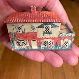 May include: A miniature two-storey house model made of printed metal, featuring a red tiled roof, a brick-patterned lower facade, cream-coloured upper walls, multiple lattice-style windows with shutters, a central door, and a small side terrace.
