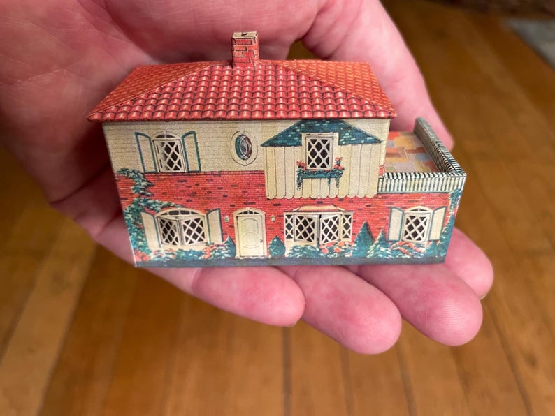 May include: A miniature two-storey house model made of printed metal, featuring a red tiled roof, a brick-patterned lower facade, cream-coloured upper walls, multiple lattice-style windows with shutters, a central door, and a small side terrace.