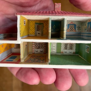 May include: A miniature two-storey dollhouse cross-section featuring a red roof and multiple rooms with printed interior details. The walls display various wallpaper patterns, including yellow, green, and blue designs, with depicted windows, doors, and rugs.