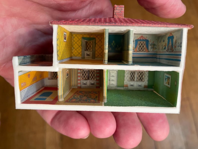 May include: A miniature two-storey dollhouse cross-section featuring a red roof and multiple rooms with printed interior details. The walls display various wallpaper patterns, including yellow, green, and blue designs, with depicted windows, doors, and rugs.