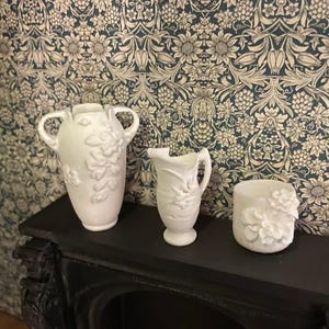 以下が含まれることがあります： 白いセラミック製の装飾品3点。花柄のレリーフ模様が施されています。取っ手付きの花瓶、小さなピッチャー、円筒形の容器が含まれます。黒い表面に配置され、花柄の壁紙を背景にしています。