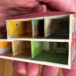 May include: A miniature two-storey dollhouse structure with an open front, featuring a red roof with a brick chimney. The interior walls are decorated with various patterns including yellow diamond, green, and striped wallpaper, with printed floor details.