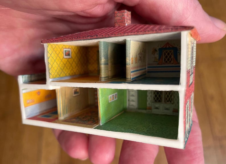 May include: A miniature two-storey dollhouse structure with an open front, featuring a red roof with a brick chimney. The interior walls are decorated with various patterns including yellow diamond, green, and striped wallpaper, with printed floor details.