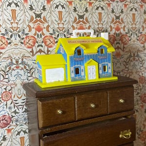May include: A miniature toy house with a yellow roof and accents, blue walls, and a white garage. The house has a white handle and the words "PLAY FAMILY HOUSE" printed on the roof. It sits on a dark brown wooden dresser.
