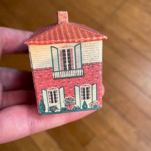 May include: A small, rectangular, house-shaped tin container featuring a printed design of a two-storey building with a red tiled roof, a brick lower level, a cream-coloured upper level, green shutters, and small bushes along the base.