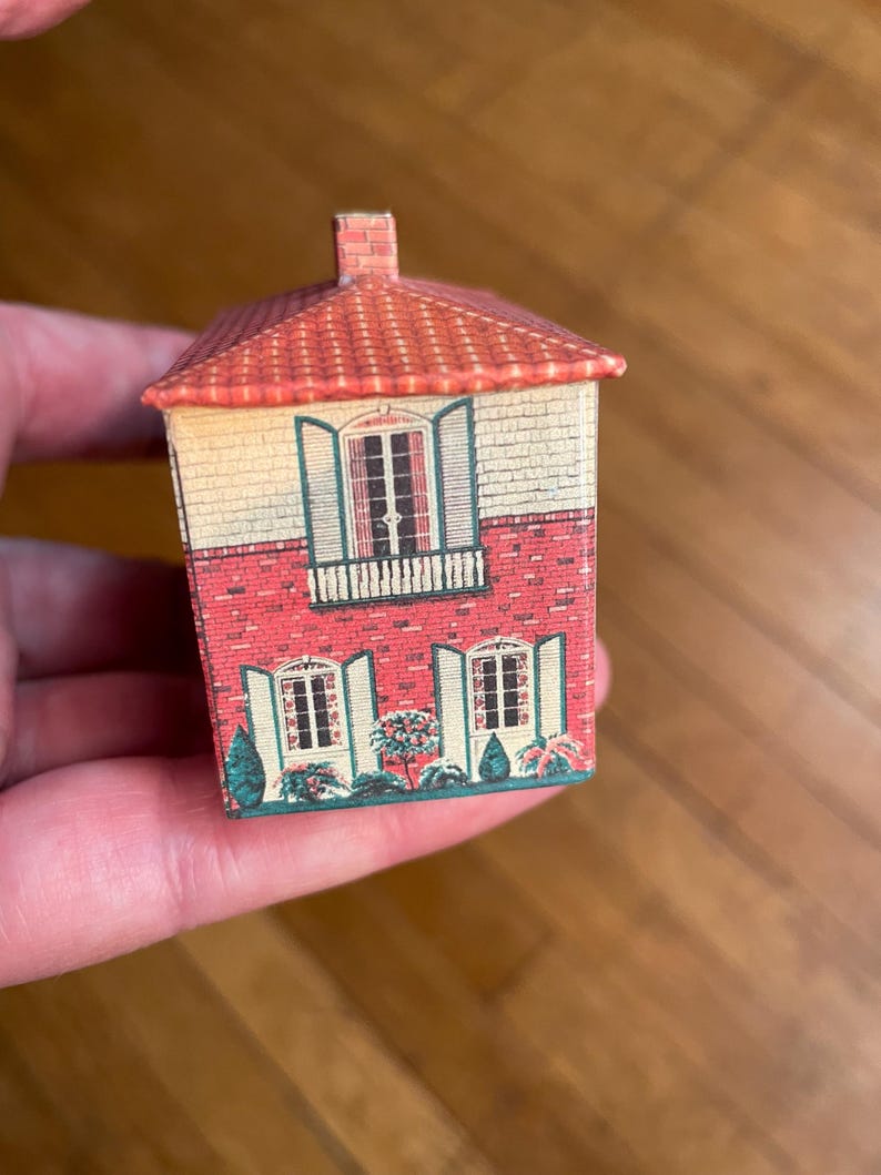 May include: A small, rectangular, house-shaped tin container featuring a printed design of a two-storey building with a red tiled roof, a brick lower level, a cream-coloured upper level, green shutters, and small bushes along the base.
