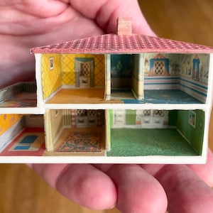 May include: A miniature dollhouse cross-section featuring two levels with multiple rooms. The structure has a red tiled roof, white exterior walls, and interior walls decorated with various patterned wallpapers, including yellow, green, and blue designs.