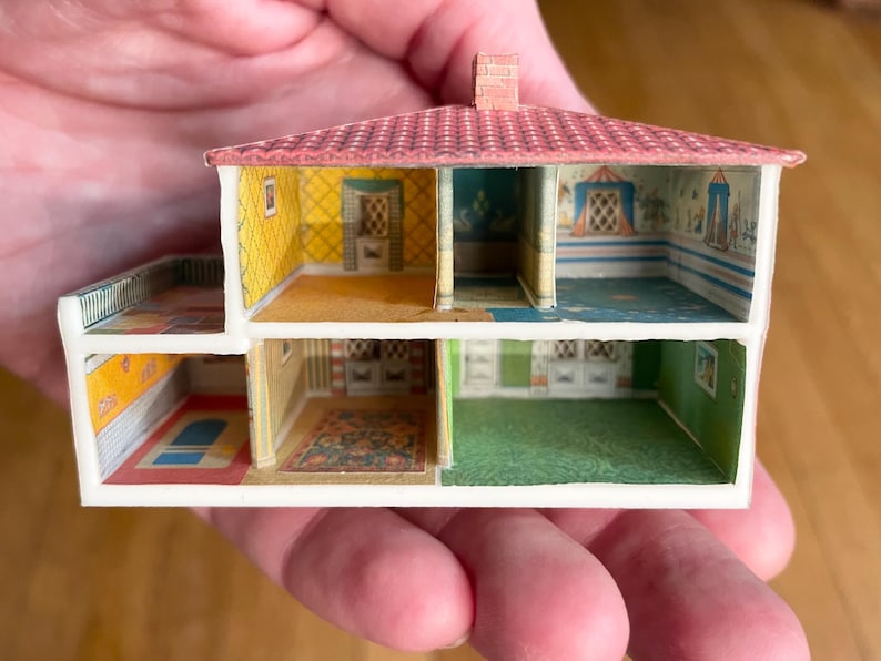 May include: A miniature dollhouse cross-section featuring two levels with multiple rooms. The structure has a red tiled roof, white exterior walls, and interior walls decorated with various patterned wallpapers, including yellow, green, and blue designs.