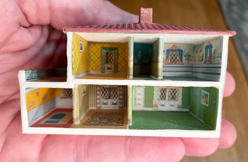 May include: A miniature two-storey dollhouse structure featuring an open-front design with multiple rooms. The interior walls display colourful patterned wallpaper in yellow, green, and blue, while the exterior roof is a textured reddish-brown.