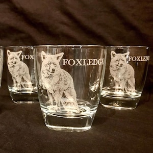 May include: Four clear glass tumblers with etched fox illustrations and the word "FOXLEDGE" in white. The fox is depicted in a standing pose. The glasses are on a dark brown surface.