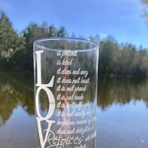 May include: A clear glass vase with the word "LOVE" etched vertically on the side. The vase is engraved with the text "Love is patient, is kind, it does not envy, it does not boast, it is not proud, it is not rude, it is not self-seeking, it is not easily angered, keeps no record of wrong, does not delight in evil, rejoices with the truth, always protects, always trusts, always hopes, always perseveres, never fails."