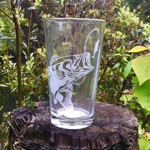 May include: A clear glass with a white etched design of a bass fish with a fishing lure in its mouth. The glass is sitting on a wooden stump.