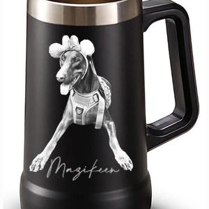 May include: Black stainless steel beer stein with a black and white image of a dog wearing a hat and harness. The dog's name, "Magifeen", is written in cursive at the bottom of the mug. The mug has a black handle and a silver rim.