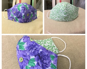 May include: Three face masks. One mask features a purple floral pattern with green leaves and yellow centers. Another mask has a green and white leaf pattern. The third mask shows both patterns.