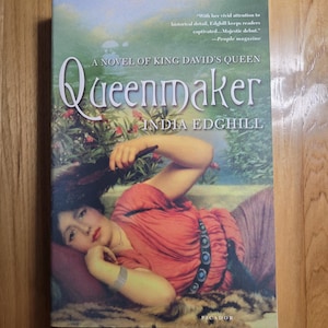 May include: A book cover for "Queenmaker" by India Edghill. The cover features a woman in a red dress reclining on a patterned surface. The title and author's name are prominently displayed, with a quote from People magazine.