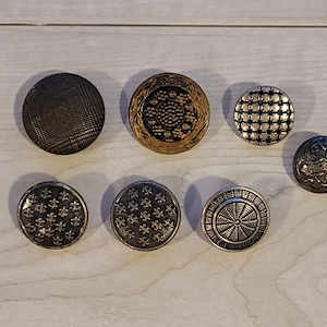 May include: Seven decorative buttons with various designs. The buttons are round and have a metallic finish. Some buttons have a floral design, while others have geometric patterns.