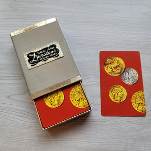 Puede incluir: Una caja vintage de cartas de juego Durastone con revestimiento de plástico y un interior rojo. La caja está abierta y muestra una baraja de cartas con monedas de oro en la cara de cada carta. Las monedas muestran imágenes de figuras históricas y el año 1905.