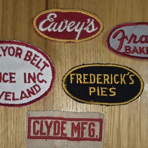 May include: A collection of vintage embroidered patches in various shapes. The patches feature text such as "Eavey's", "Frantz Bakers", "Conveyor Belt Service Inc. Cleveland", "Frederick's Pies", and "Clyde Mfg.". The patches are in red, white, black, and gold.