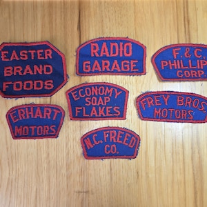 Könnte beinhalten: Eine Sammlung von Vintage-Patches in Blau und Rot mit gestickten Schriftzügen. Die Patches zeigen Firmennamen wie "Easter Brand Foods", "Radio Garage" und "F. & C. Phillips Corp." auf einer Holzoberfläche.