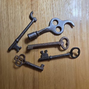 May include: Five antique keys of varying designs and sizes, made from aged metal, are displayed on a wooden surface. The keys feature different handle shapes, including a heart, a circle, and a decorative swirl. The keys' aged appearance and intricate designs are clearly visible.