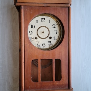 May include: A vintage wooden wall clock with a white face and black Roman numeral markings. The clock has a brass trim and a pendulum.