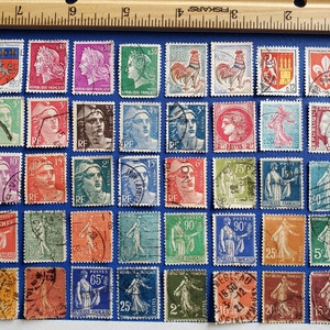 May include: A collection of vintage French postage stamps, featuring portraits, allegorical figures, and heraldic symbols. The stamps are in various colors, including red, blue, green, and purple, and are arranged on a blue surface. A wooden ruler is in the background.