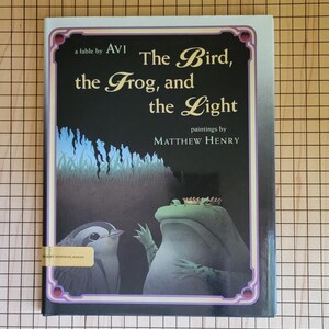 May include: A book cover with a black background and a purple border. The title is "The Bird, the Frog, and the Light" with the author listed as "AVI" and the illustrator as "MATTHEW HENRY". The cover features a green frog with a gold crown and a small grey bird.