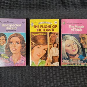 May include: Three vintage paperback books with colorful covers. Titles include "Unexpected Hazard," "The Flight of the Hawk," and "The Mouth of Truth." Each cover features illustrations of people and text, with a Harlequin Romance logo.