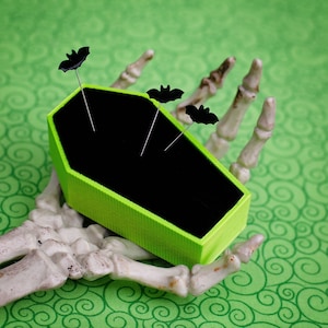 May include: A lime green coffin-shaped pincushion with three black bat pins inserted. The pincushion is held by a skeletal hand.