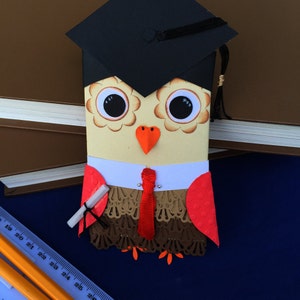 May include: A graduation card featuring an owl wearing a graduation cap and holding a diploma. The owl is brown, white, and red with black eyes. The card is decorated with lace and a red bow tie.