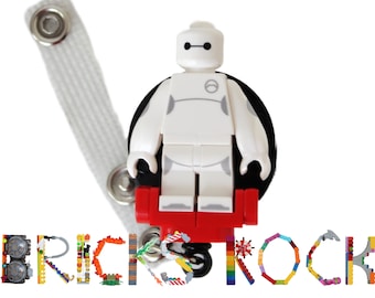May include: White Lego minifigure of Baymax from the Disney movie Big Hero 6. The minifigure is attached to a black and red Lego badge reel. The badge reel is attached to a white strap. The word "BRICKSROCK" is spelled out in Lego bricks below the minifigure.