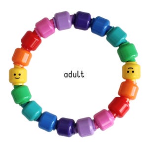 Op de afbeelding: Een kleurrijke armband gemaakt van speelgoedbouwstenen in een regenboog van kleuren. De armband bevat twee gele blokken met lachende gezichten. Het woord "adult" is in zwarte tekst gedrukt.