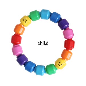 Op de afbeelding: Een kleurrijke armband gemaakt van regenboogkleurige blokken, elk met een lachend gezicht. Het woord "child" staat op de afbeelding. Een vrolijk accessoire.