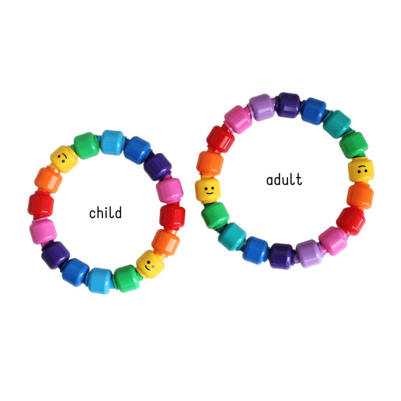 Op de afbeelding: Twee regenboogkleurige kralenarmbanden, &eacute;&eacute;n met het label "kind" en de andere "volwassene". Elke armband heeft een reeks kleurrijke kralen, waaronder rood, oranje, geel, groen, blauw, paars en roze. Sommige kralen hebben lachende gezichten.