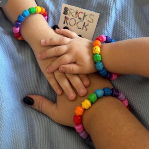 Op de afbeelding: Twee regenboogkleurige kralenarmbanden, &eacute;&eacute;n om de pols van een volwassene en &eacute;&eacute;n om de pols van een kind. De armbanden zijn gemaakt van kleurrijke blokken. Een kaart met de tekst "BRICKS KOCK" is zichtbaar.
