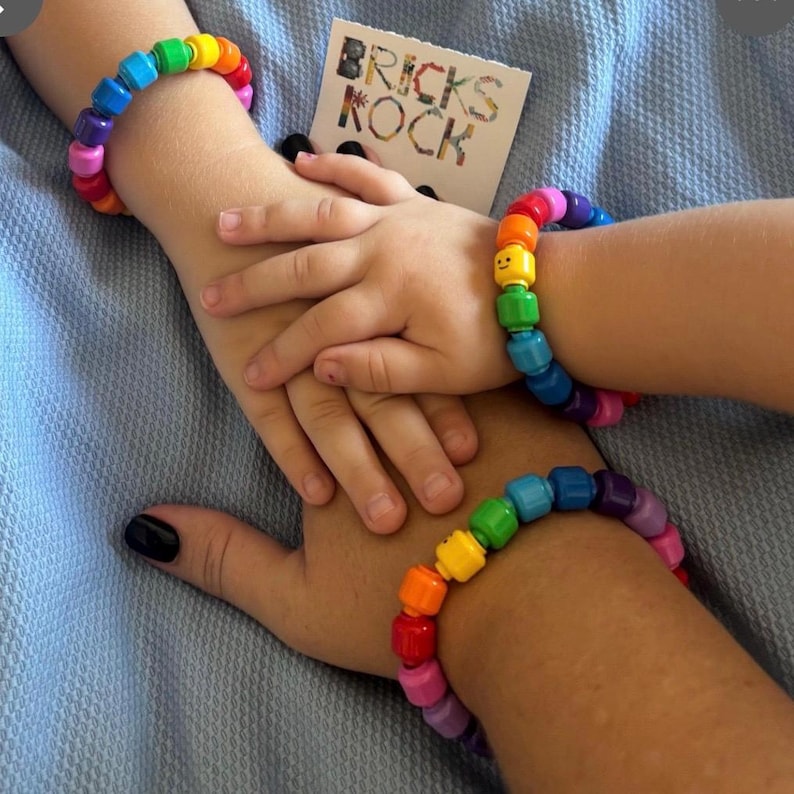 Op de afbeelding: Twee regenboogkleurige kralenarmbanden, &eacute;&eacute;n om de pols van een volwassene en &eacute;&eacute;n om de pols van een kind. De armbanden zijn gemaakt van kleurrijke blokken. Een kaart met de tekst "BRICKS KOCK" is zichtbaar.
