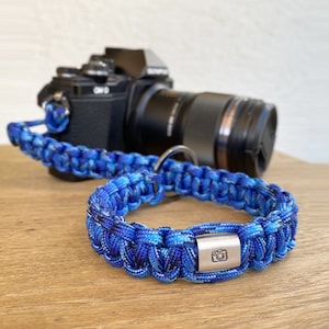 Könnte beinhalten: Ein blaues und schwarzes Kameraband aus Paracord mit einem silbernen Kamerareiz. Das Band ist an einer schwarzen Kamera mit einem silbernen Objektiv befestigt.