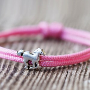 Schmales Kinderarmband aus rosa Nylonseil. Durch Knoten in der Größe verstellbar. Versilberter Schieber zum Beispiel ein Pferd. 
Weitere Schieber: Herz vsilber, Herz vergoldet, versilberte Hundepfote, Buddha versilbert, Kommunionsfisch versilbert