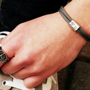 Könnte beinhalten: Ein silberner Ring mit Kompass-Design und ein graues Armband mit silbernem Akzent, das eine Abschlusskappe und die Zahl 20 zeigt. Das Armband besteht aus einer dunkelgrauen Kordel.